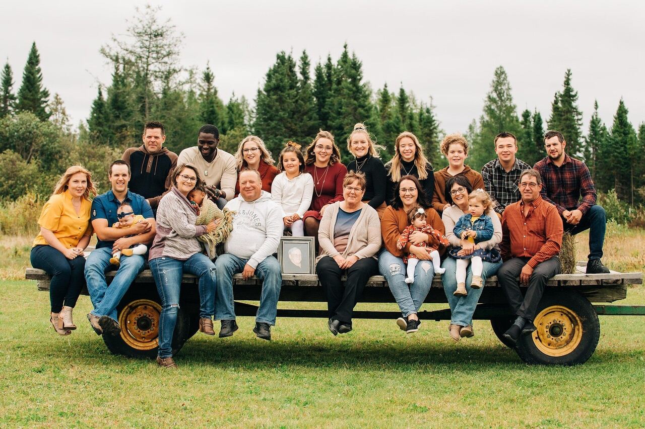 A large family of three generations sitting together on a farm trailer in a field, with pine trees behind
