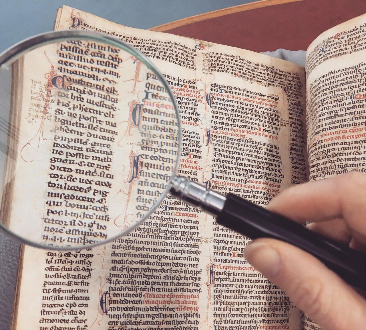 A hand holding a magnifying glass over the handwritten text of an old illuminated manuscript